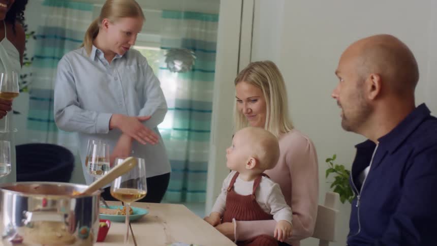 A group of people stand and sit at a table with a woman holding a baby talking after dinner