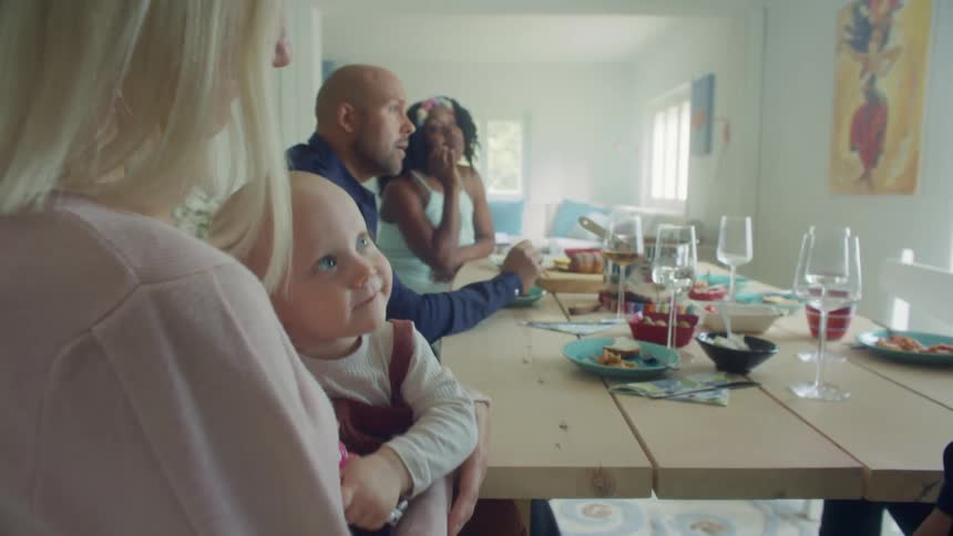 A woman holding a baby at a table in a room with people in the background