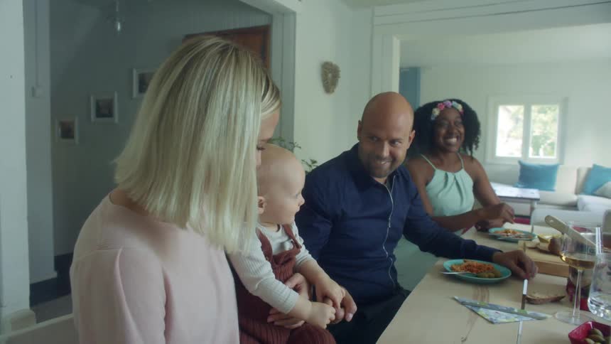 A woman holds a baby sitting at a home dinner table with another man and woman having conversation