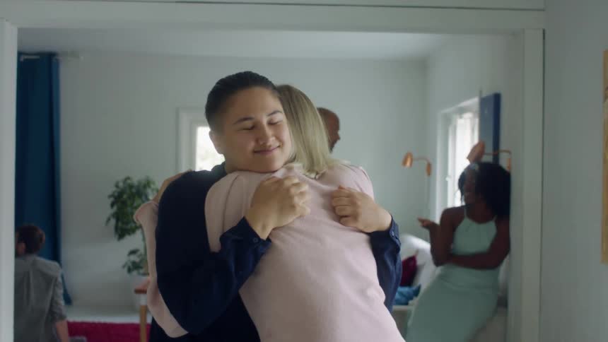 Two woman hug in front of a man a woman chatting while standing in a dining room