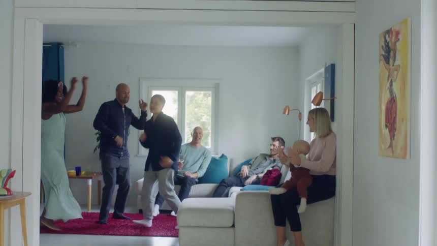 People dance in the middle of the living room as others watch and clap sitting on the couch