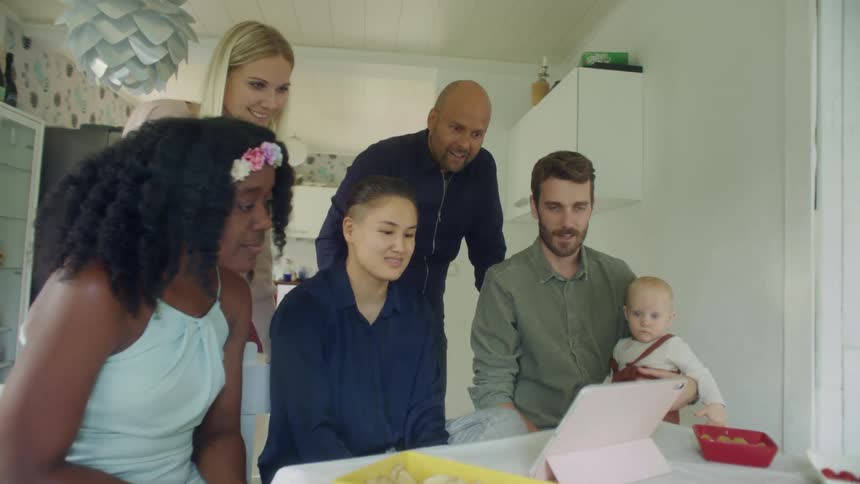 A group of people speak with one another while sitting around a computer tablet on a stand on a dining table