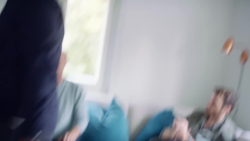 A man dances with another person in a small room with others sitting on a couch