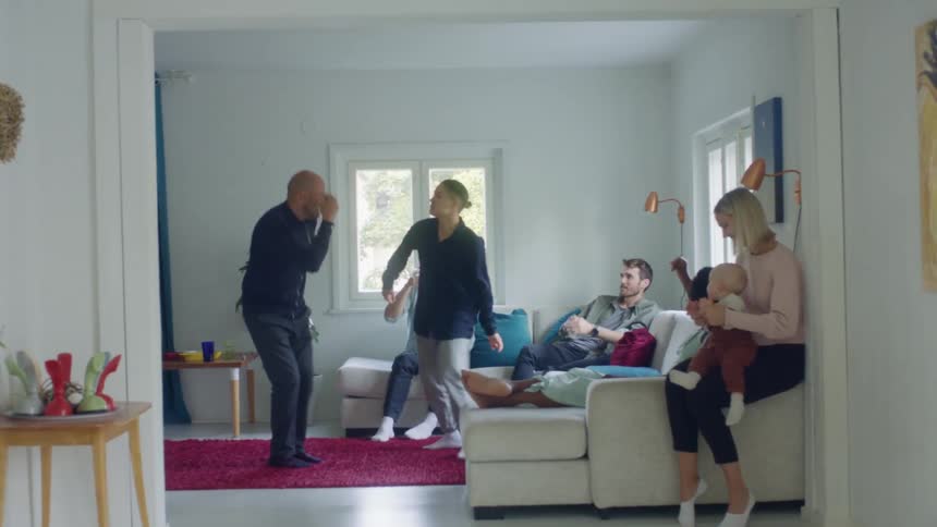 Two people dance together in a living room with other people sitting on the couch watching