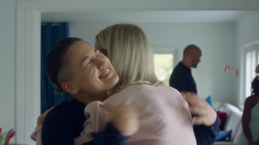 Two women hug excitedly as their friends hang out in the living room behind them