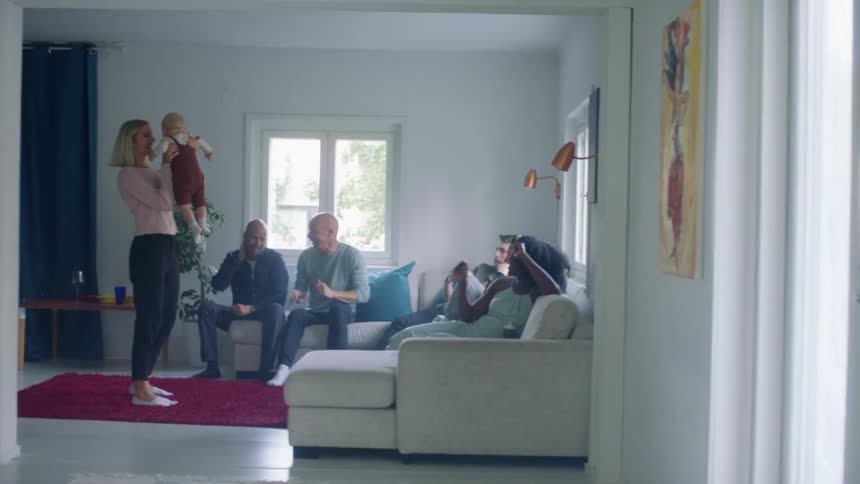 A woman dances with her baby in a living room while her friends cheer her on while sitting on the couch in a living room