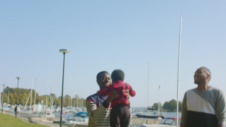 A male toddler is picked up by his dad as they stand next to boats and a body of water