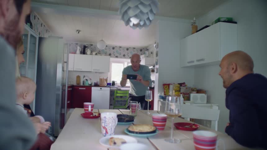 A man uses his smartphone to a take photograph of a group of people sitting at a dining table in a home dining room during the day