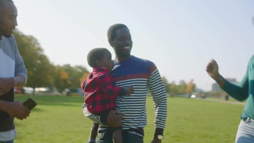A man holds his toddler as he and two friends stand in a semi circle and dance in a park