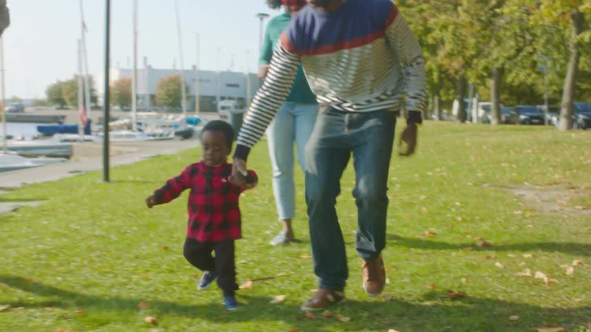 A group of adults run alongside a toddler through a field bordering a dock during the day