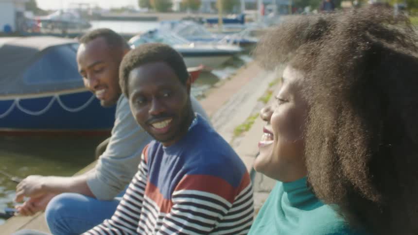 Two men and a woman talk and laugh while sitting on the wooden docks on a lake during the day