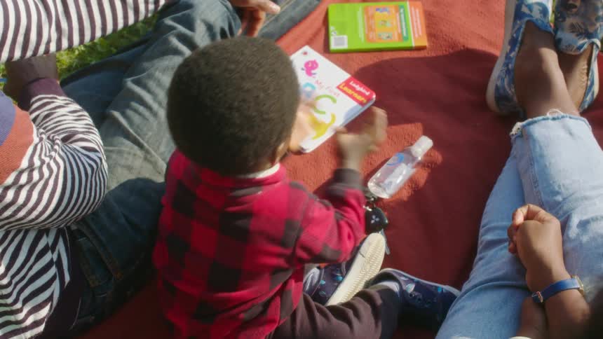 A toddler uses hand sanitizer and grabs a book to read while sitting on a blanket in a park with this parents