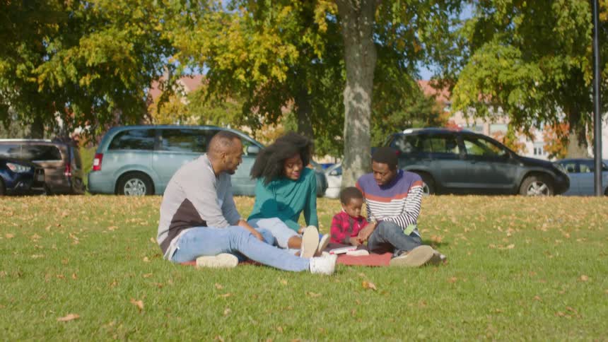 A family sits on a blanket in the park and reads to their toddler son