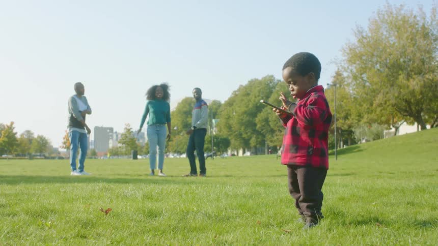 Toddler with a smartphone dances on the grass of a park in the city with his family
