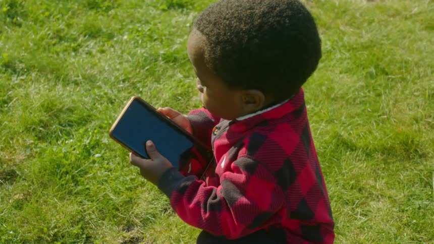 Toddler black boy with a plaid shirt picks up smartphone on the grass in the public park