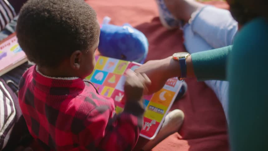 Young boy sitting next to his mother on a blanket in the grass while she teaches him to read a picture book at daytime