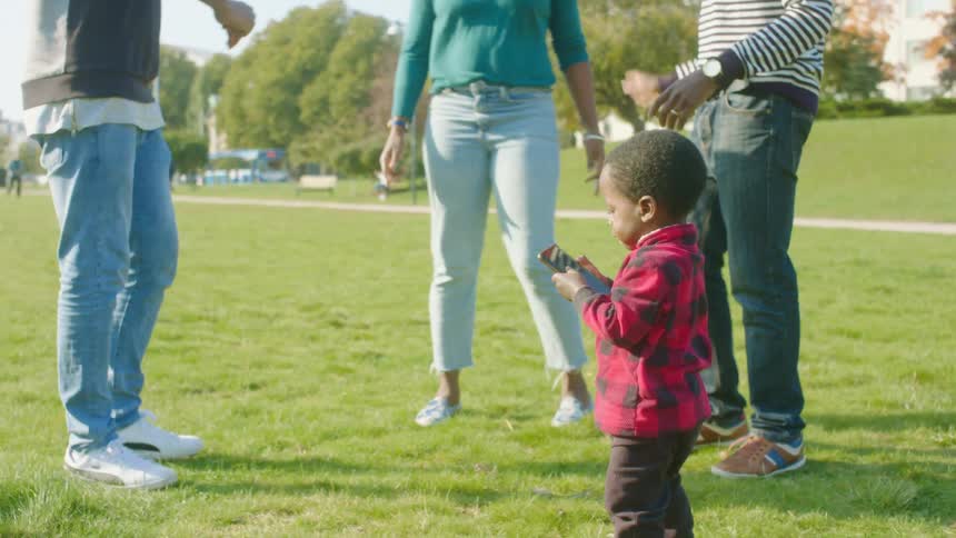 A toddler picks up a cellphone on the ground in a park as his parents dance around him