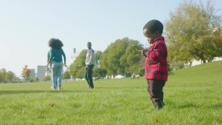 A small boy dances while playing with a smartphone in a field as a group of adults dance nearby
