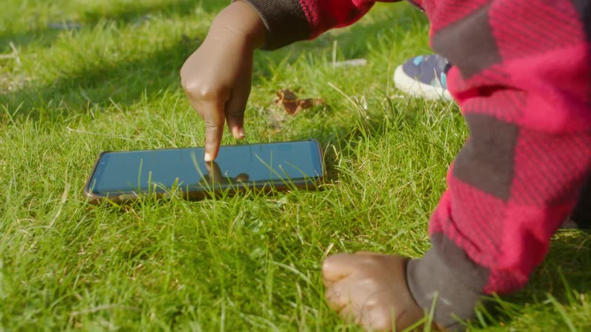 Black toddler playing with a cell phone on the lawn at a park