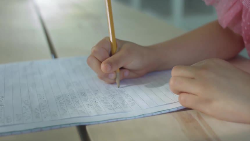 A young girl uses a pencil to write in a notebook while sitting at a wooden table