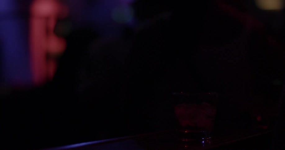 A woman stands in front of a bar counter with an alcoholic drink as lights flash around her