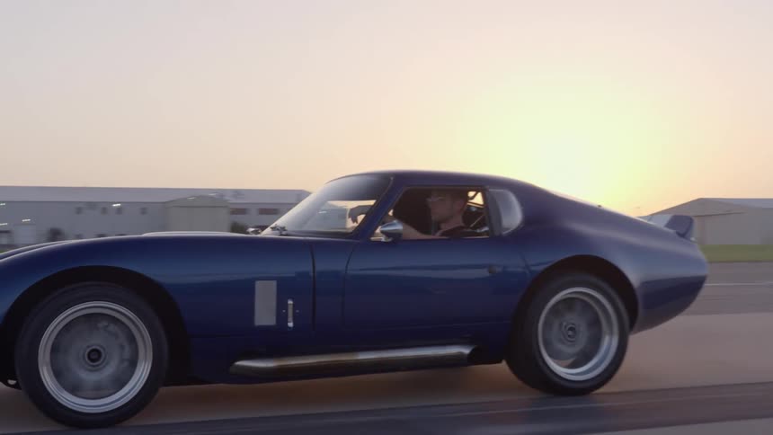 A man drives a blue sports car down an airstrip in the evening