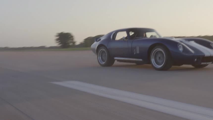 A man drives a blue vintage sports car through a parking lot next to a field in the countryside