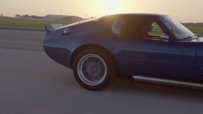 A man drives a blue vintage sports car down a road past a field in the countryside