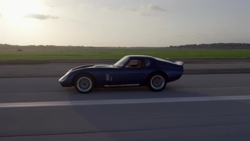 A blue sports car driving on a rural paved road on a summer day