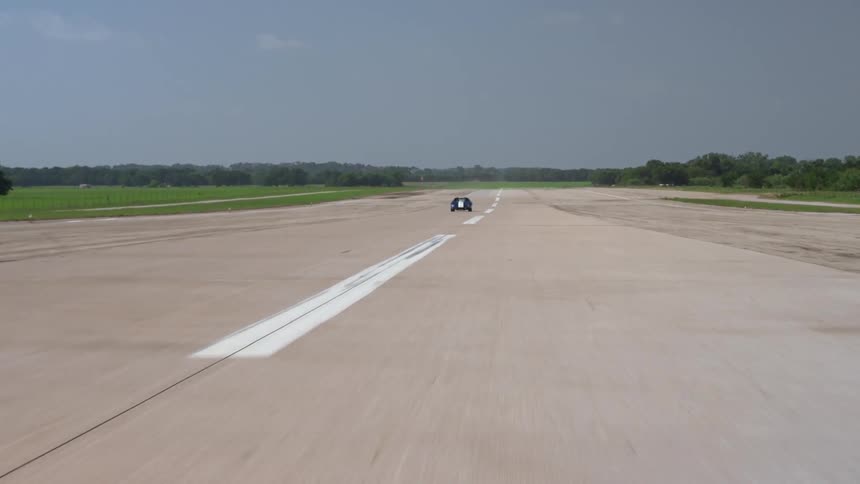 Sports car speeds down wide road between fields bordered by forest