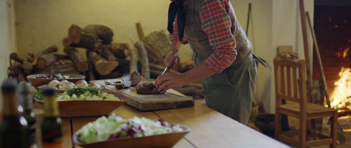 A person stands at a table cutting food on a cutting board in a room with a fire burning in a fireplace