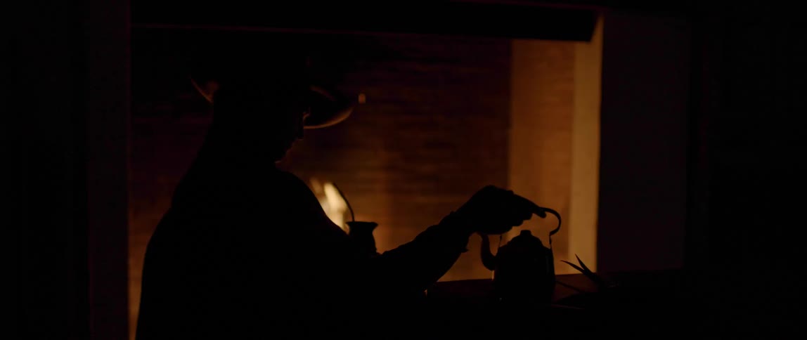 Silhouette of a man pours hot water from a kettle to prepare yerba mate in front of the fireplace