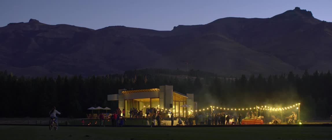 A young girl rides her bicycle toward a large house party in front of a mountain