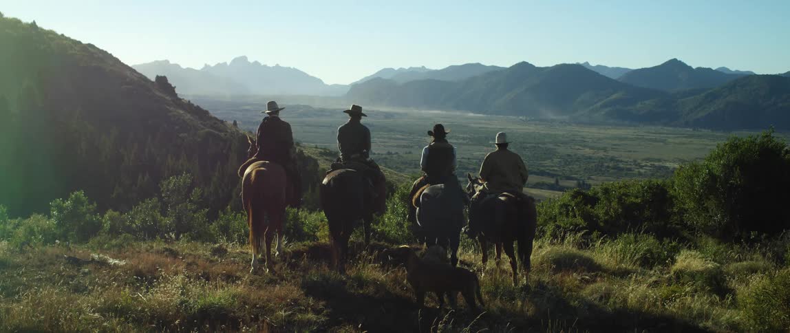 A group of cowboys on horses stand on top of a hill in front of a valley between the mountains
