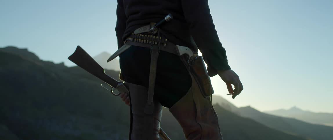 Man armed with several weapons and wearing chaps stands as sun brightens fog over mountains