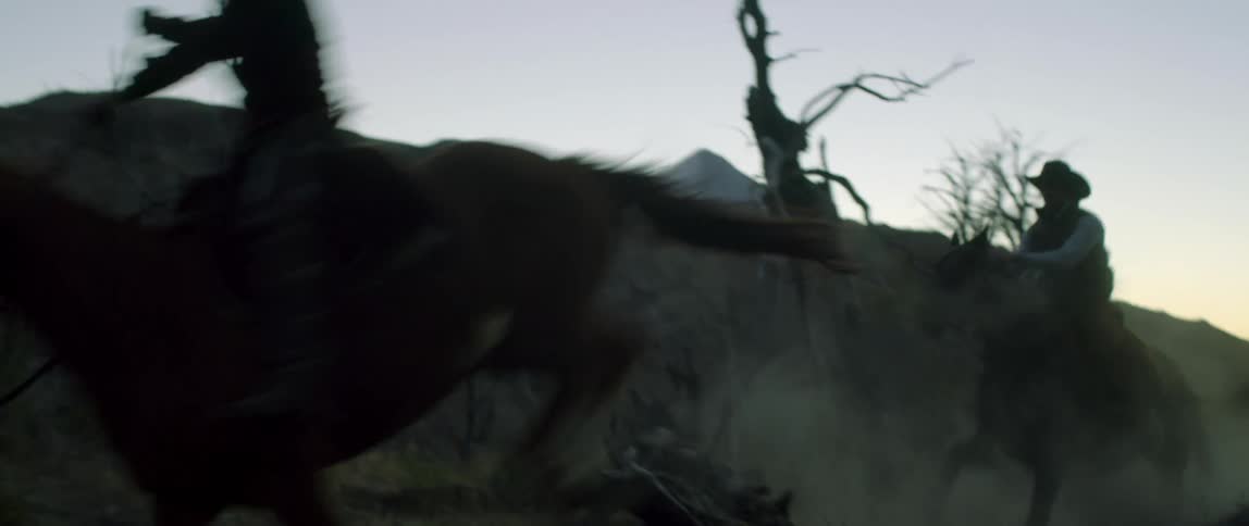 Cowboys riding horses jump dead trees in the desert next to the hills at sunset