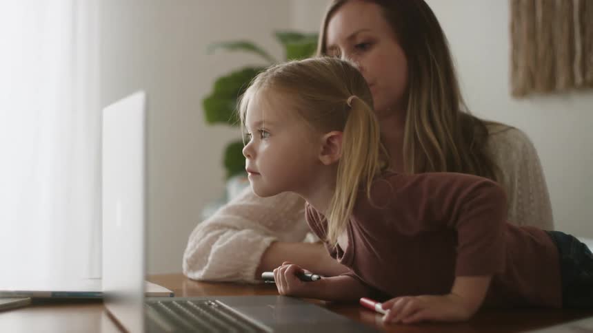 A young girl leans over a table and pushes a key on a laptop with her mother sitting beside her