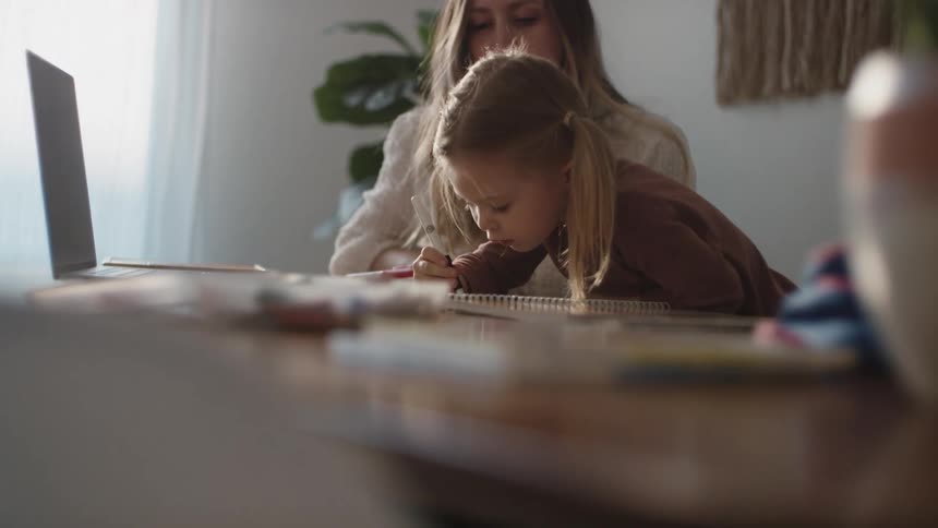 A young girl uses a marker to write in a notebook as her mother watches from beside her