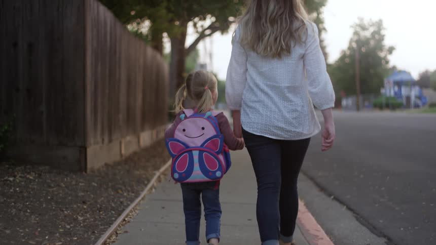 Mother and daughter walk hand in hand along sidewalk in neighborhood