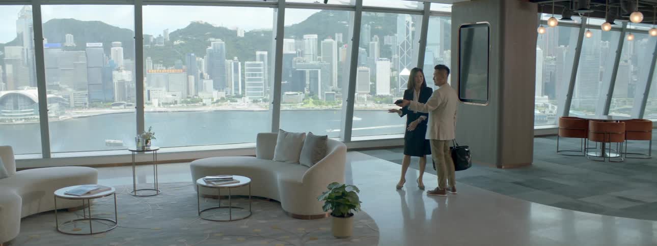 A man and a woman standing in a room with couches and tables with a large window in the background