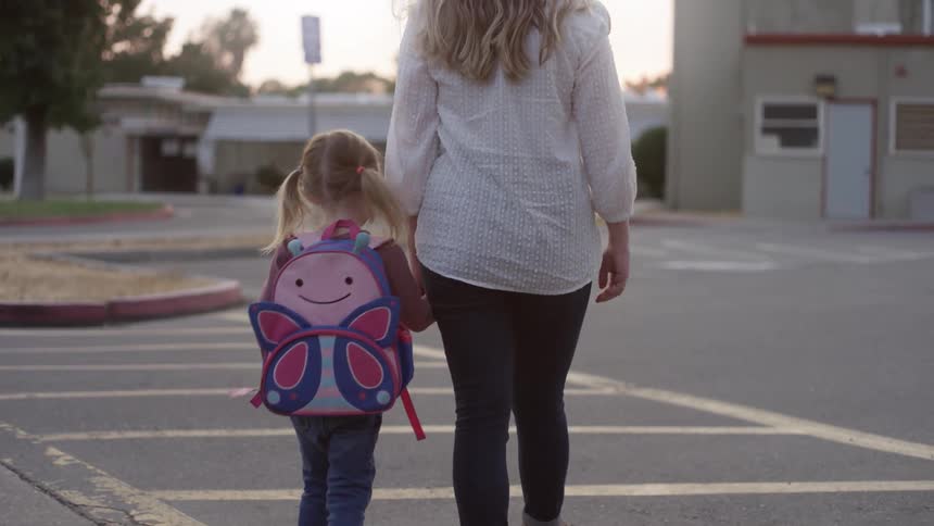 A mom and daughter hold hands as they walk through a parking lot in a neighborhood at dusk