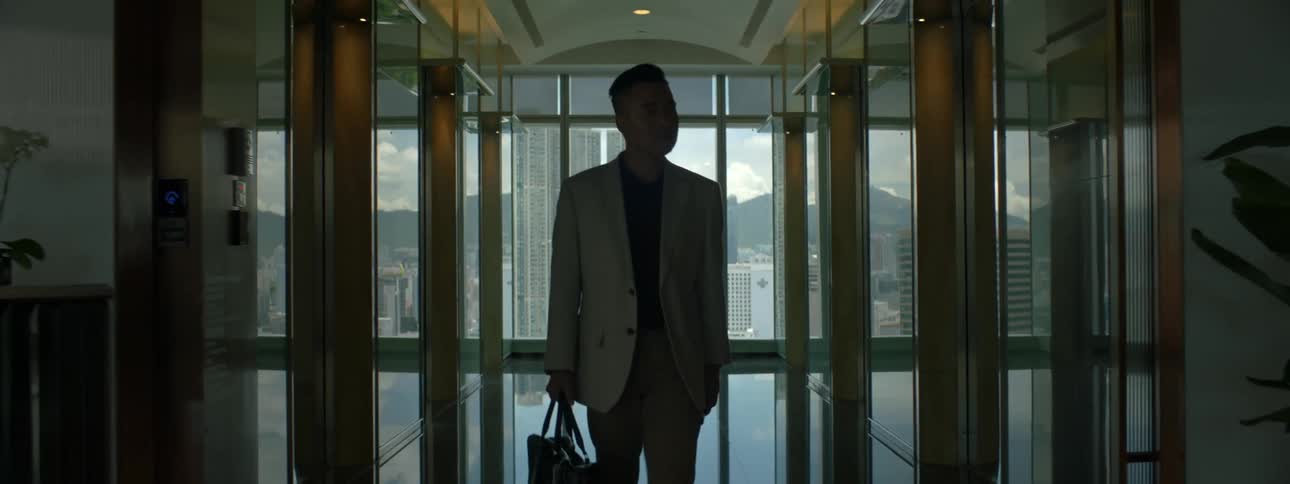 A man walking in an office building is greeted by a woman who leads him to another room