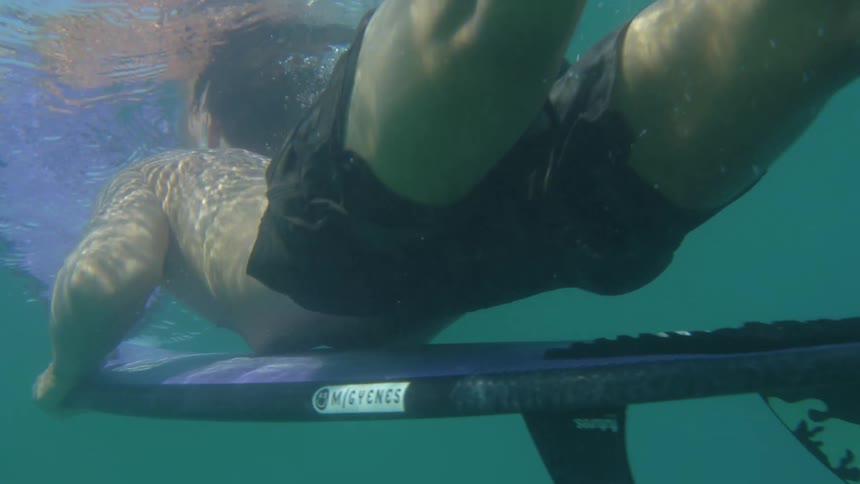 A surfer floating underwater brings his surfboard towards his chest as he floats towards the surface