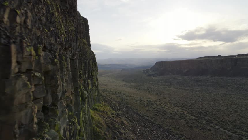 Flying closely alongside a rugged cliff wall above a wide and barren canyon valley