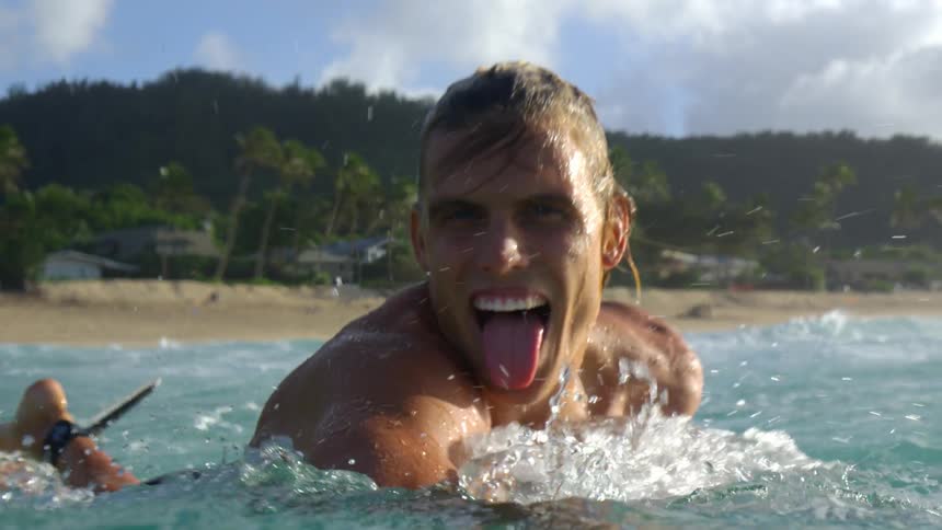 A surfer paddles on his board in the ocean while sticking his tongue out at the camera
