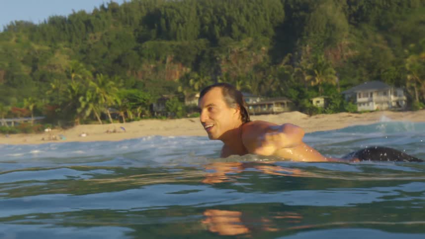 A man is paddling on a surfboard in the water smiling with a shoreline and beach behind him during the day