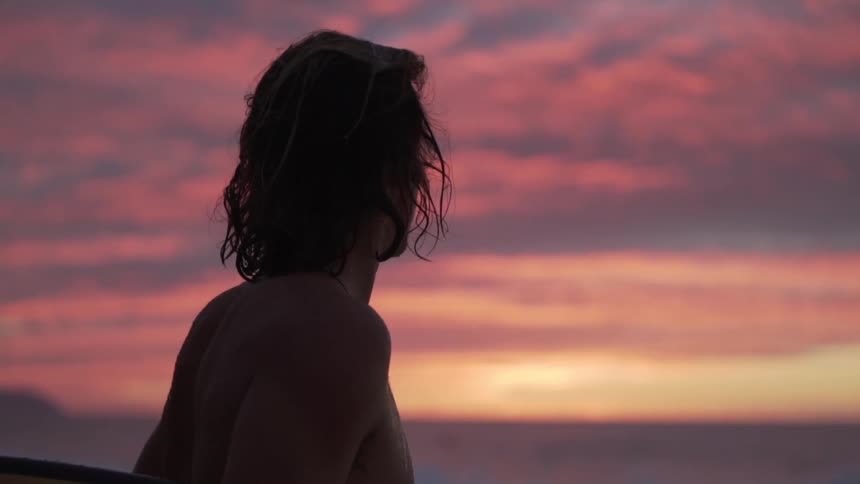 Male surfer holding a surfboard watches sea and red sky at dusk