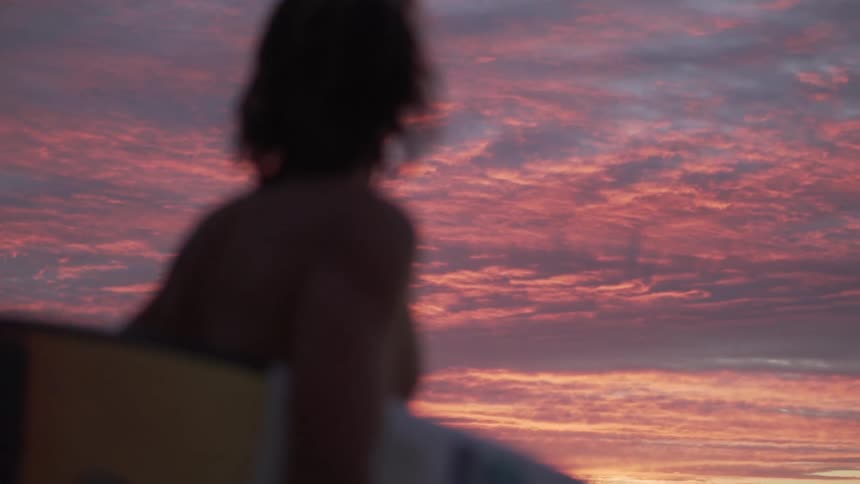 A person walks outside at sunset carrying a surf board