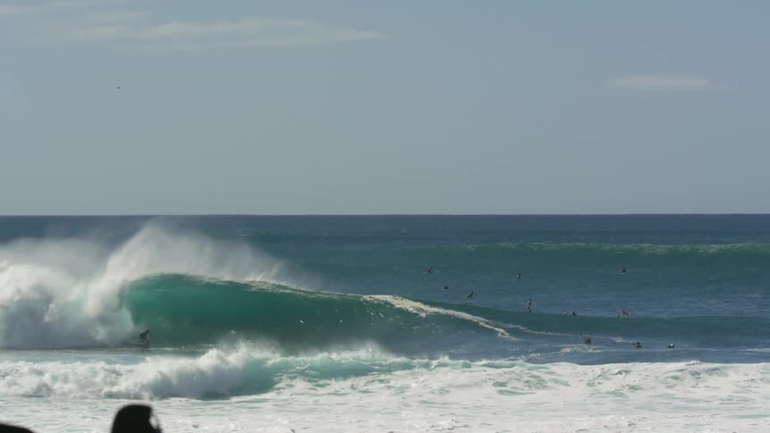 Two surfers ride a wave as other people float in the water close by and the waves go on to crash on the beach on a sunny day