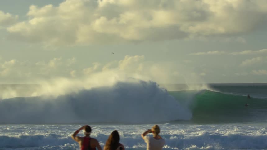 Surfer rides a surfboard under a wave while people watch on the coast at sunset